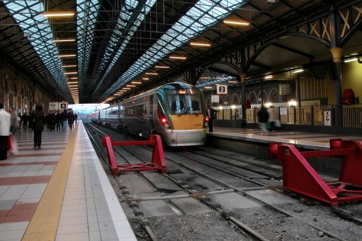 Modern train station platform with passengers boarding a sleek train under a large, arched glass roof.