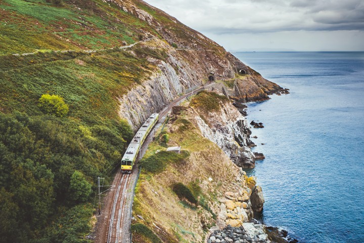 Train traveling along a scenic coastal cliff with lush greenery and ocean views under a cloudy sky.